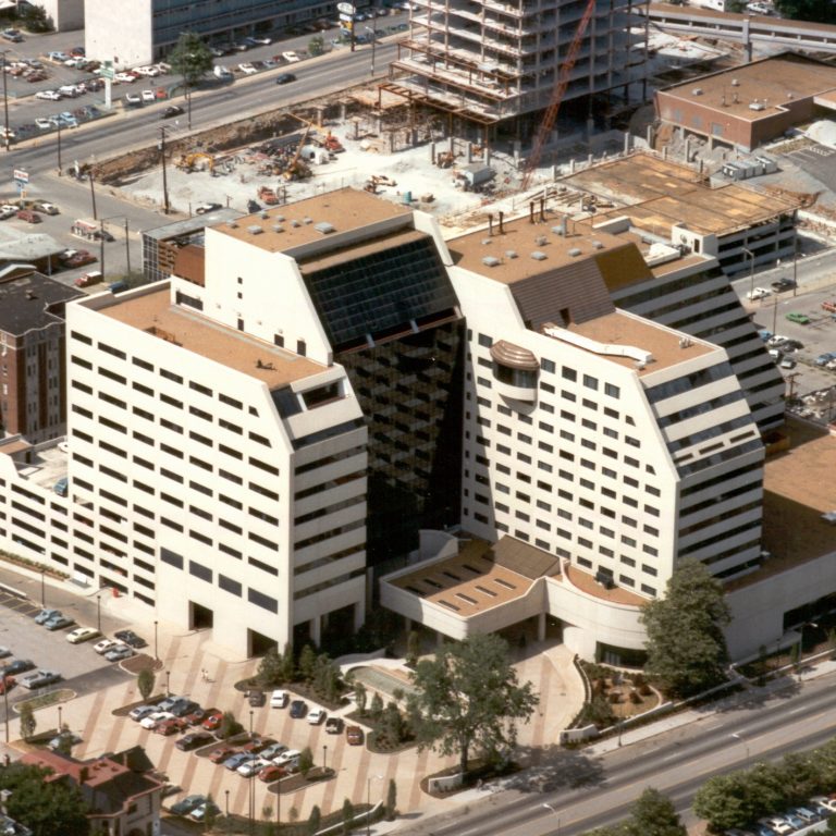 Aerial view of a modern, multi-story office building complex with angular architecture, surrounded by roads, trees, and construction sites in an urban area.