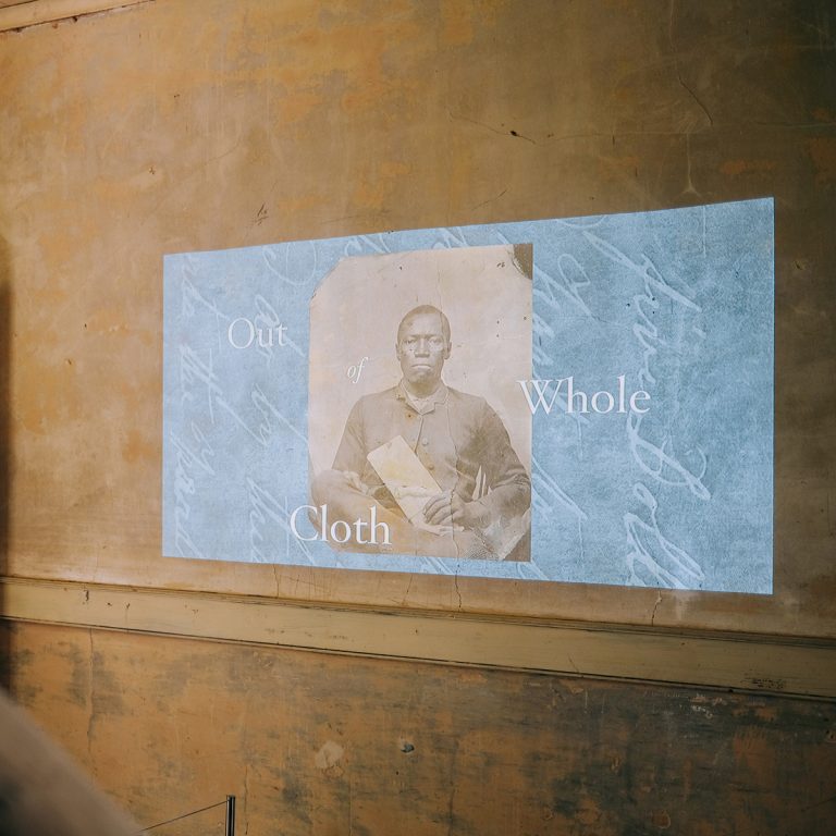 A vintage photo of a seated person with the words Out of Whole Cloth is projected onto a worn, beige wall near a staircase and an open door.