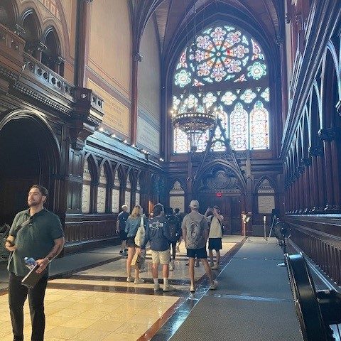 People walk inside a grand hall with high vaulted ceilings, dark wood paneling, and a large stained glass window at the far end.