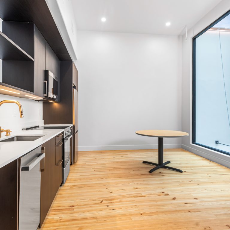 Modern kitchen with dark cabinets, white countertops, built-in appliances, a gold faucet, and a round table next to large floor-to-ceiling windows on light wood flooring.