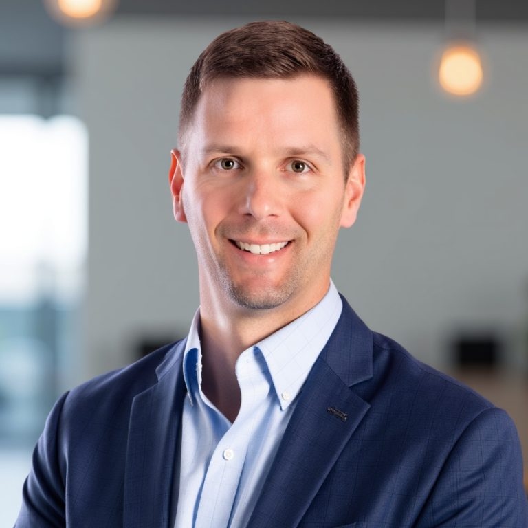 A man in a blue suit jacket and light blue shirt stands indoors, smiling at the camera with a blurred office background.