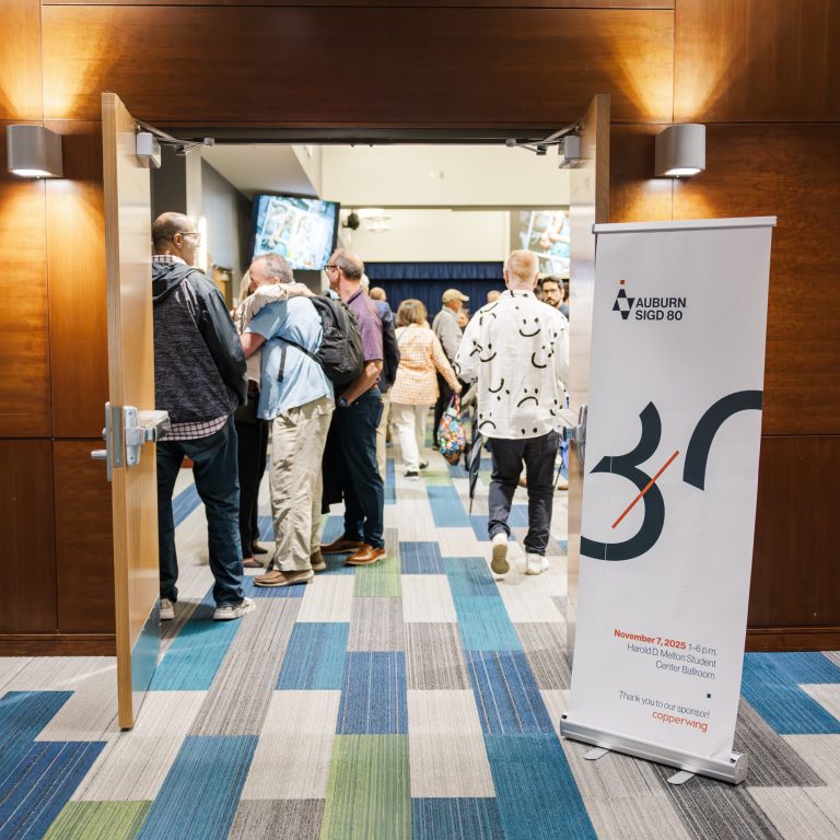 People enter a conference room through double doors; a vertical sign for the Auburn SBDC event stands to the right, and the floor has a blue and green patterned carpet.