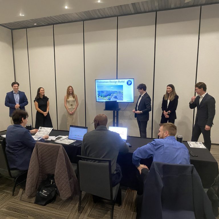 A group of six people stands presenting in front of a screen to four seated individuals in a conference room setting.