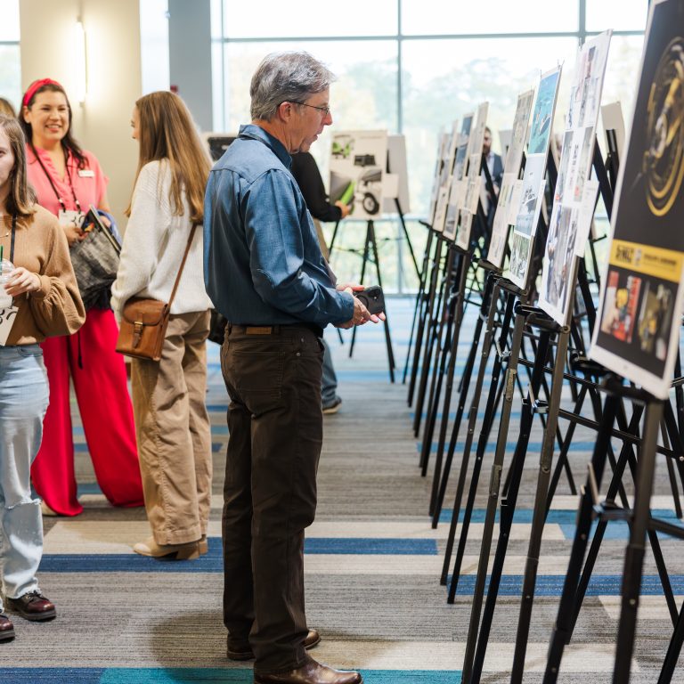 People view artworks displayed on easels in a well-lit indoor space with large windows and blue-striped carpet.