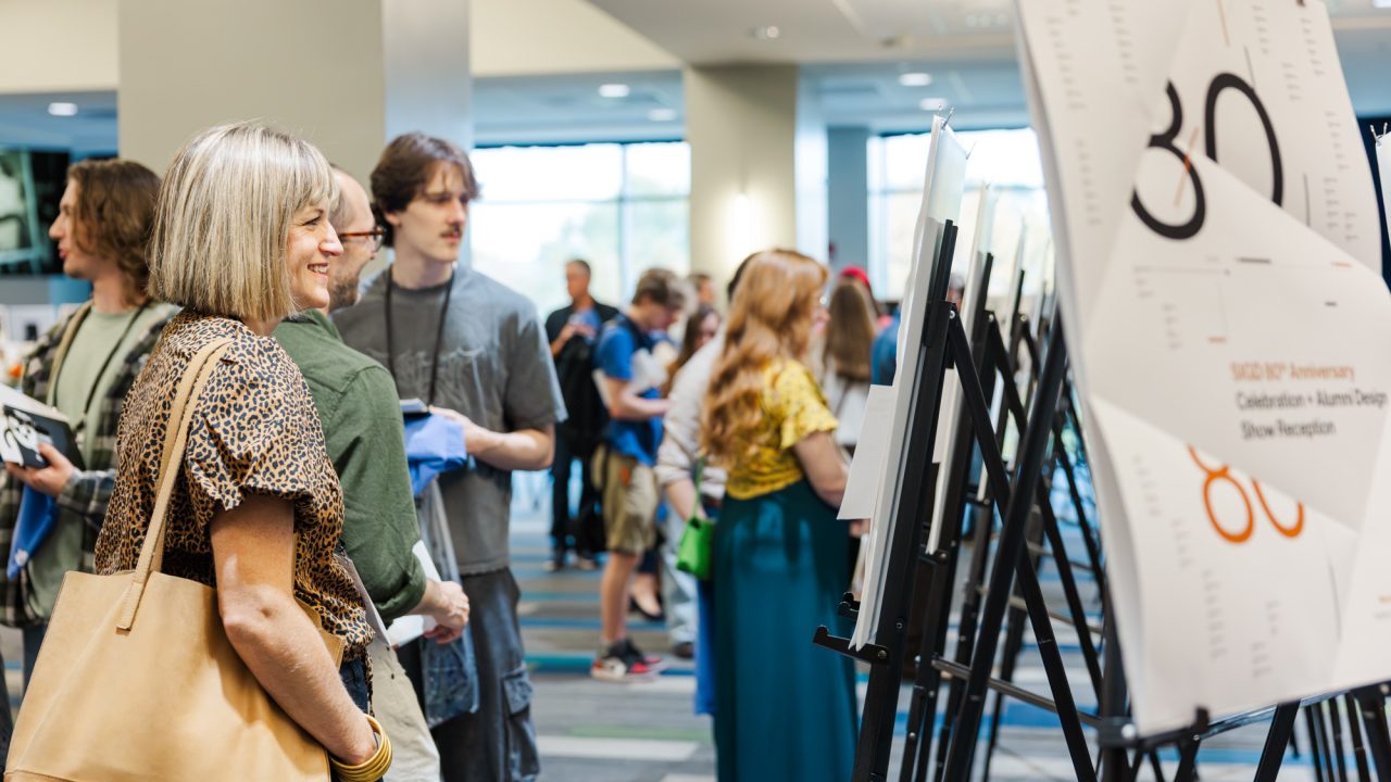 People view posters on easels at an indoor event with several attendees in the background. One sign reads 80th Anniversary Celebration + Alumni Design Show Reception.
