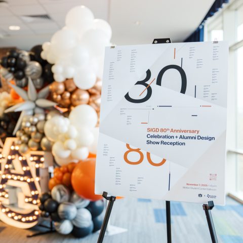 A large sign announces the SIGD 80th Anniversary Celebration and Alumni Design Show Reception, with decorative balloons and illuminated letters in the background.