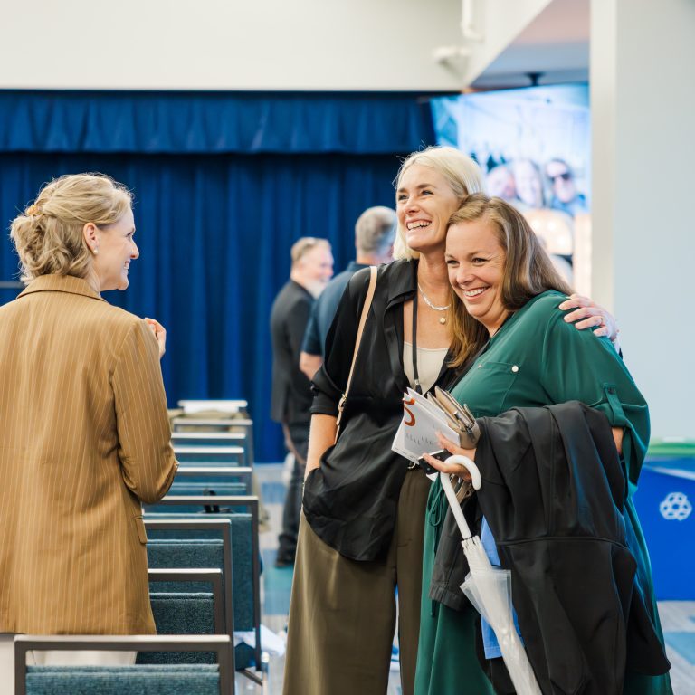 Three women stand and talk in a conference room, smiling and holding items like a coat, tote bag, and umbrella. Rows of chairs and a blue curtain are visible in the background.