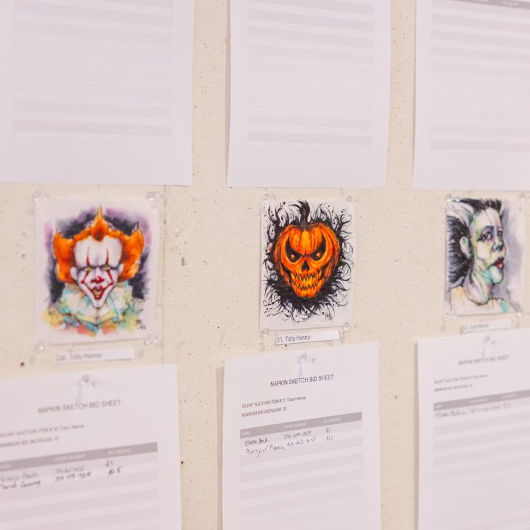 Sheets of paper and three colorful drawings of horror-inspired faces, including a clown, a pumpkin, and a pale figure, are pinned to a wall.