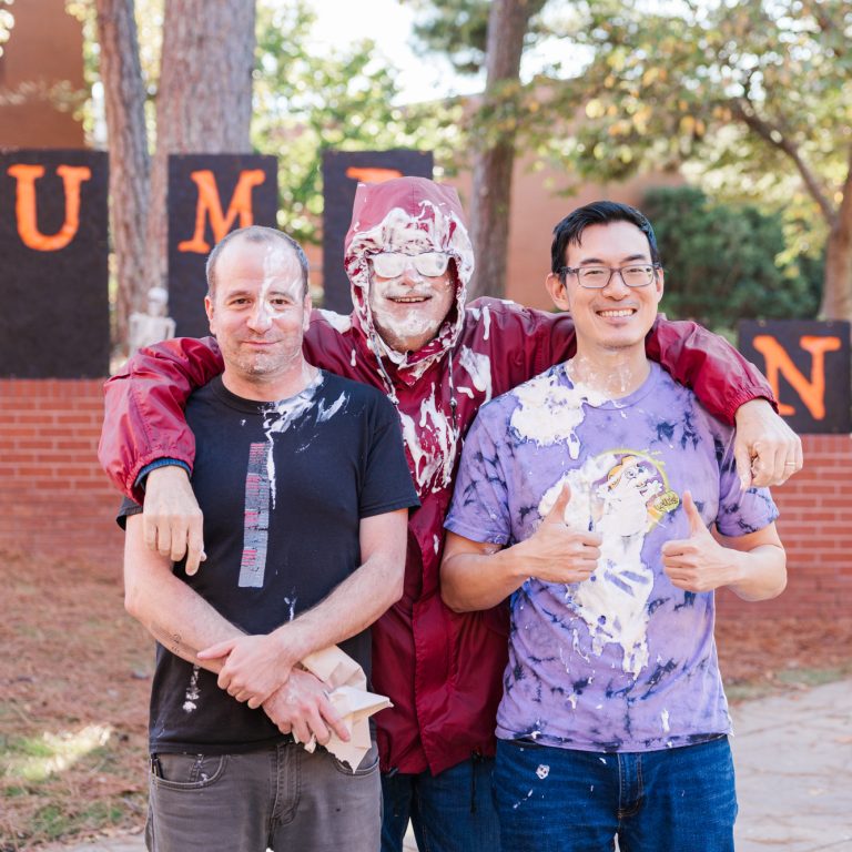 Three men stand smiling with their arms around each other, two covered in white splatter, in front of a PUMPKINS sign outdoors.