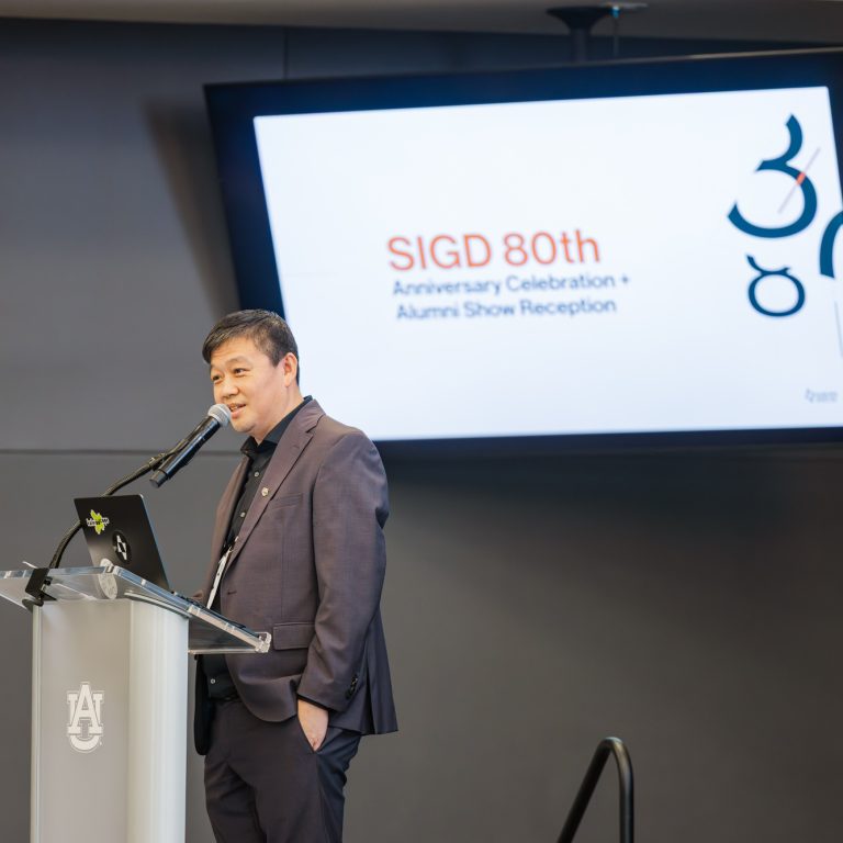 A man in a suit speaks at a podium, with a screen behind him displaying SIGD 80th Anniversary Celebration + Alumni Show Reception.