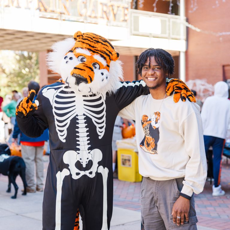 A person stands next to someone in a tiger mascot costume wearing a skeleton outfit at an outdoor event with people and carved pumpkins in the background.