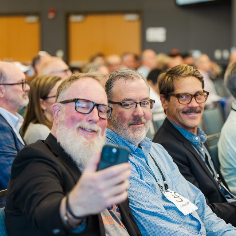 A group of men sitting in rows at a conference, with one man in front taking a selfie on his phone.