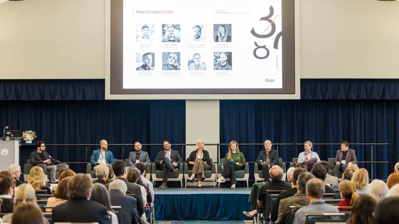 A panel of eight people sit on stage in front of an audience, with a presentation slide displaying speaker photos and text projected above them.