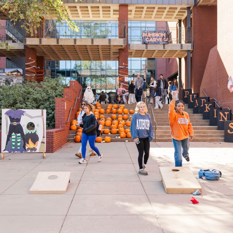 Three people play cornhole near a pumpkin display at an outdoor autumn festival; others walk and gather near decorated signs and stairs labeled PUMPKINS.