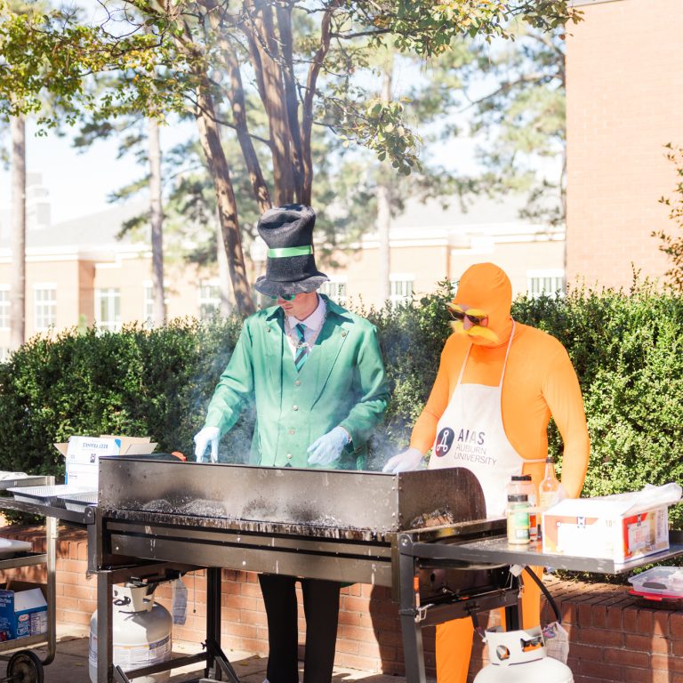 Two people in costumes are grilling food outdoors at a cookout event, with other people standing nearby and trees in the background.