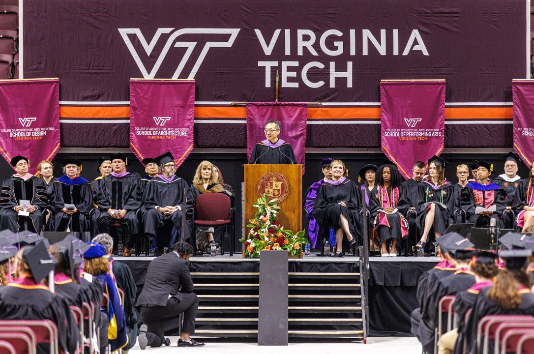 A speaker addresses graduates from a podium on stage at a Virginia Tech commencement ceremony, with faculty seated behind under school banners.