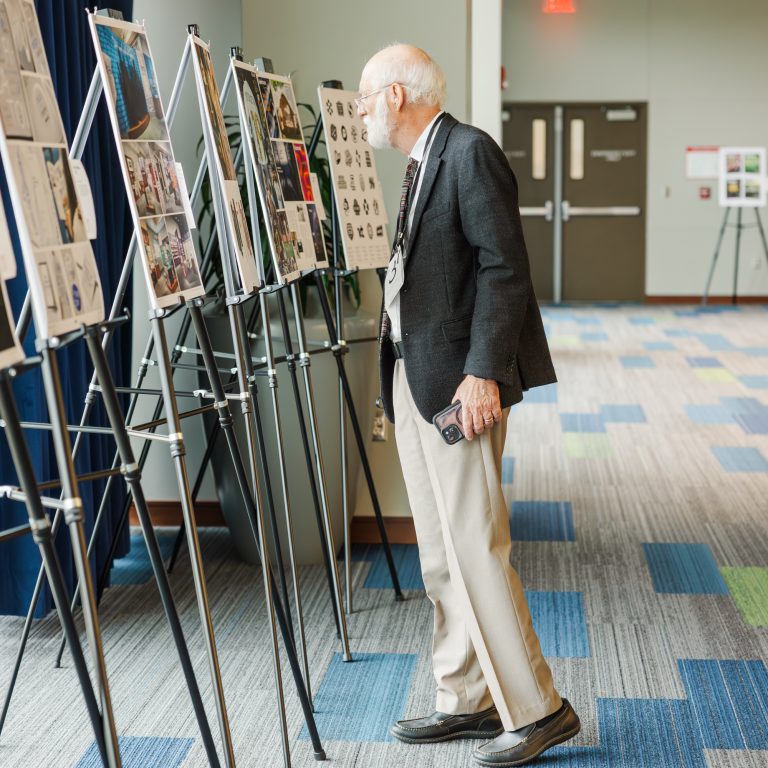 An older man in a suit jacket and khakis closely examines artwork or posters displayed on easels in a brightly lit hallway.