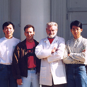 Four men stand side by side in front of a column; one wears a white lab coat, while the others wear casual clothing.