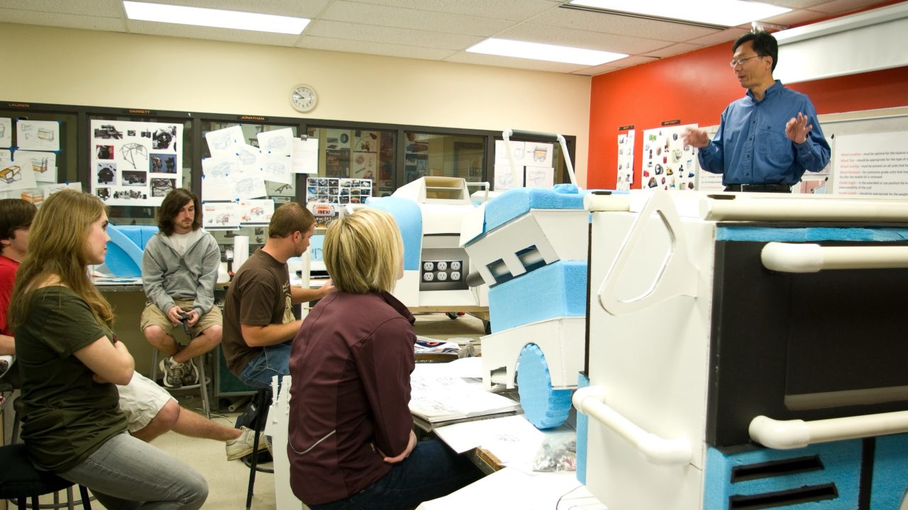 A teacher stands at the front of a classroom speaking to students seated around projects and models, with design sketches on the walls.