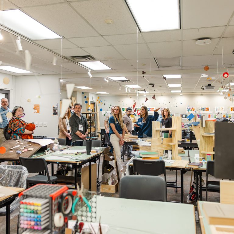 A group of people stands in a well-lit classroom filled with art supplies, student projects, and hanging decorations.
