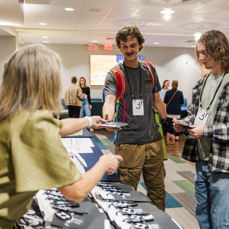 Two young men receive name tags and event materials from a woman at a registration table in a brightly lit room.
