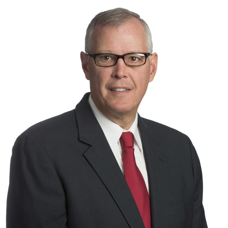A man in a dark suit, white shirt, red tie, and glasses poses against a white background.