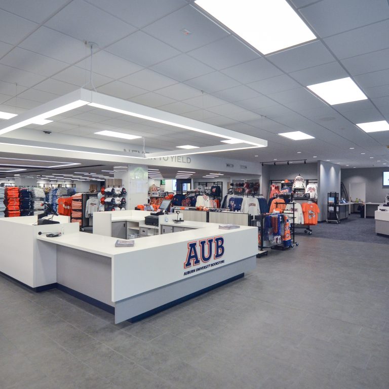 Modern university bookstore interior with merchandise displays, checkout counters, hats, clothing racks, and a sign reading AUB. Bright lighting and organized layout.