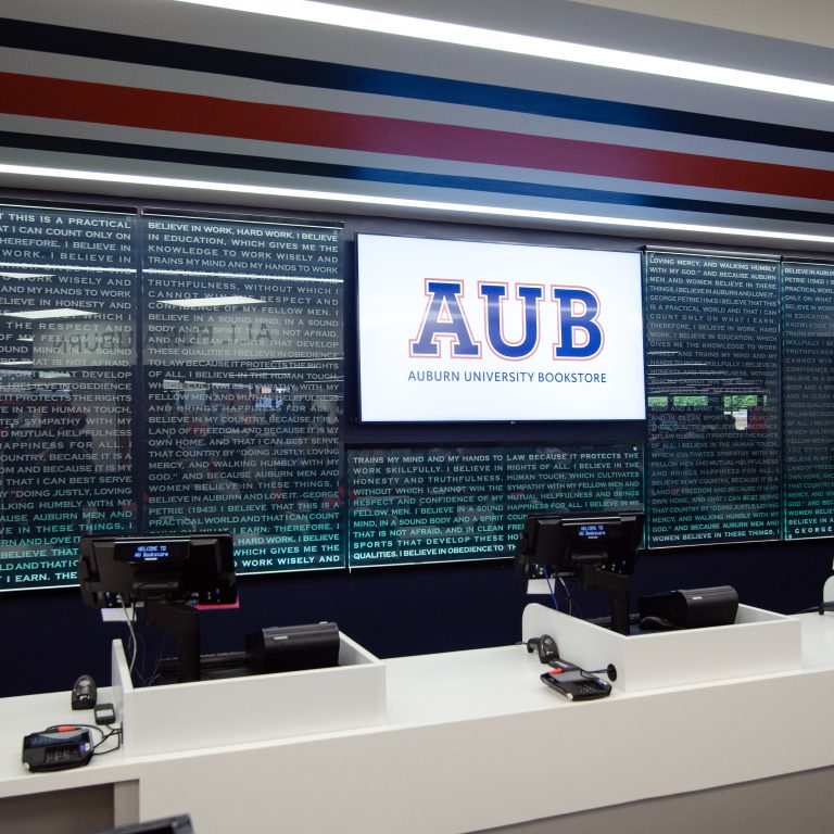 A checkout counter at the Auburn University Bookstore with electronic registers and a large digital sign displaying AUB Auburn University Bookstore on the wall.