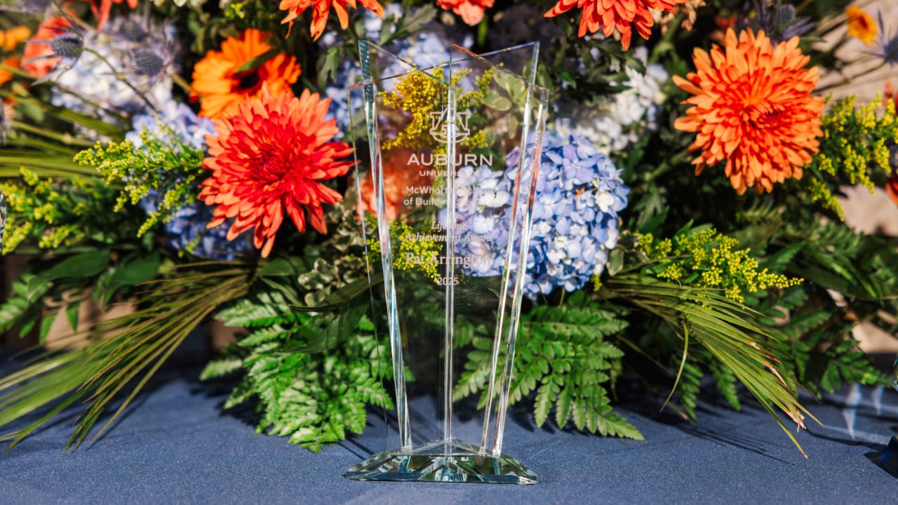 A clear glass award trophy is displayed on a blue tablecloth in front of a large floral arrangement with orange, blue, and purple flowers.