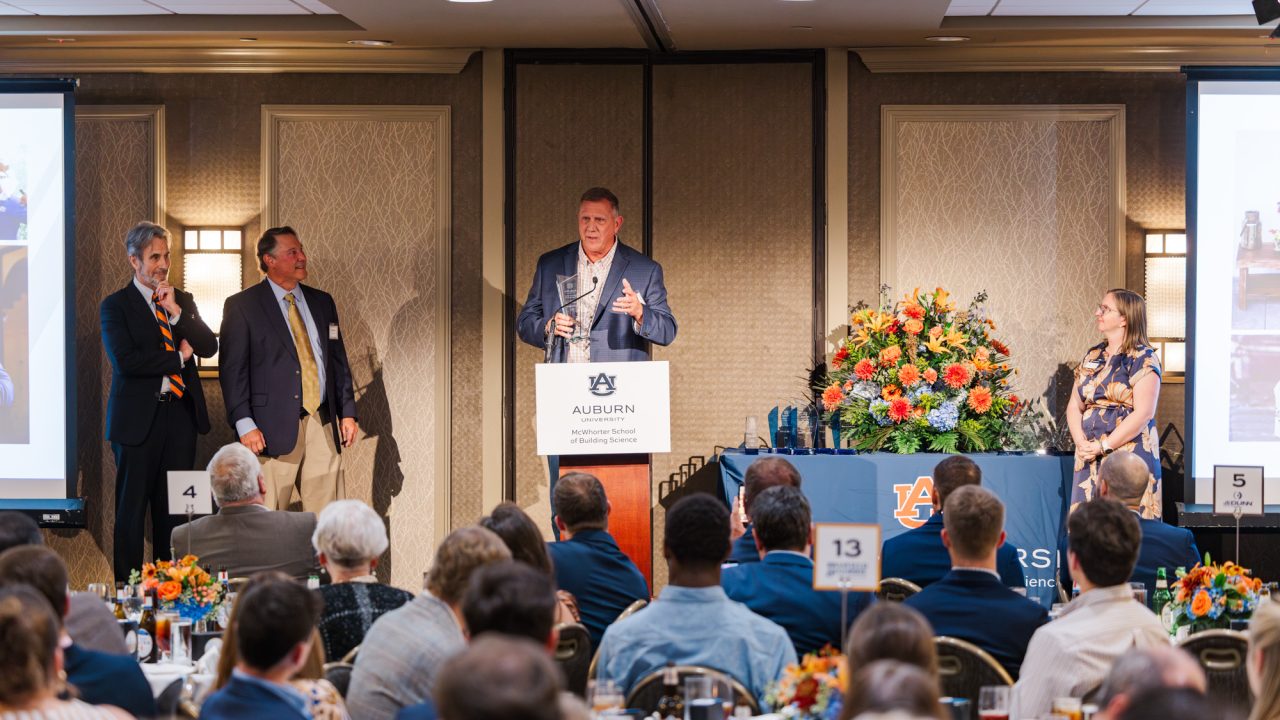 A man speaks at a podium with the Auburn University logo during an indoor event, with three people on stage and a seated audience in front.