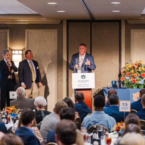 A man speaks at a podium with the Auburn University logo during an indoor event, with three people on stage and a seated audience in front.