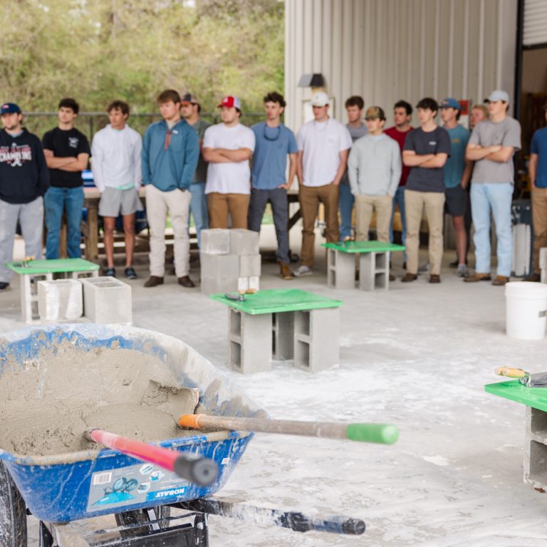 A group of people stands in a garage, observing several workstations made of cinder blocks and green boards; a wheelbarrow filled with cement is in the foreground.