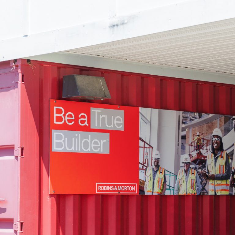 A red shipping container displays a sign reading Be a True Builder with a photo of construction workers in safety gear and the Robins & Morton logo.