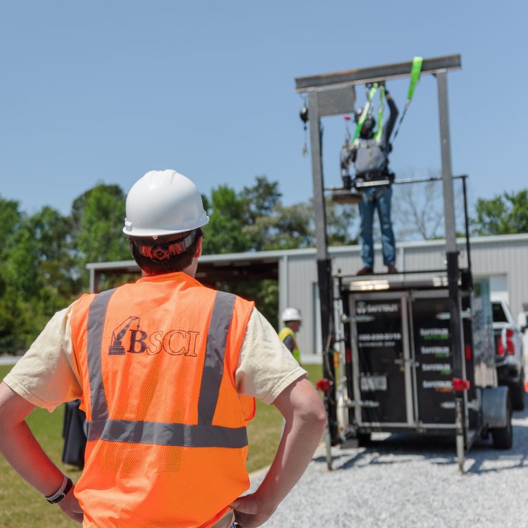 A person in an orange BSCI safety vest observes another worker using fall protection equipment on a training structure outdoors.