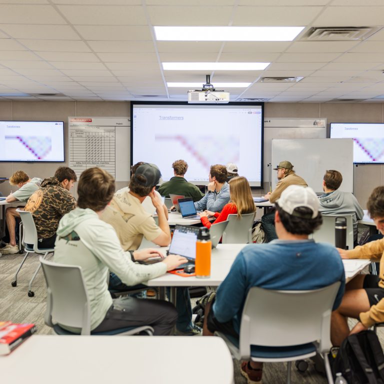 Students sit at tables in a classroom, facing a teacher and presentation slides displayed on three large screens at the front of the room.