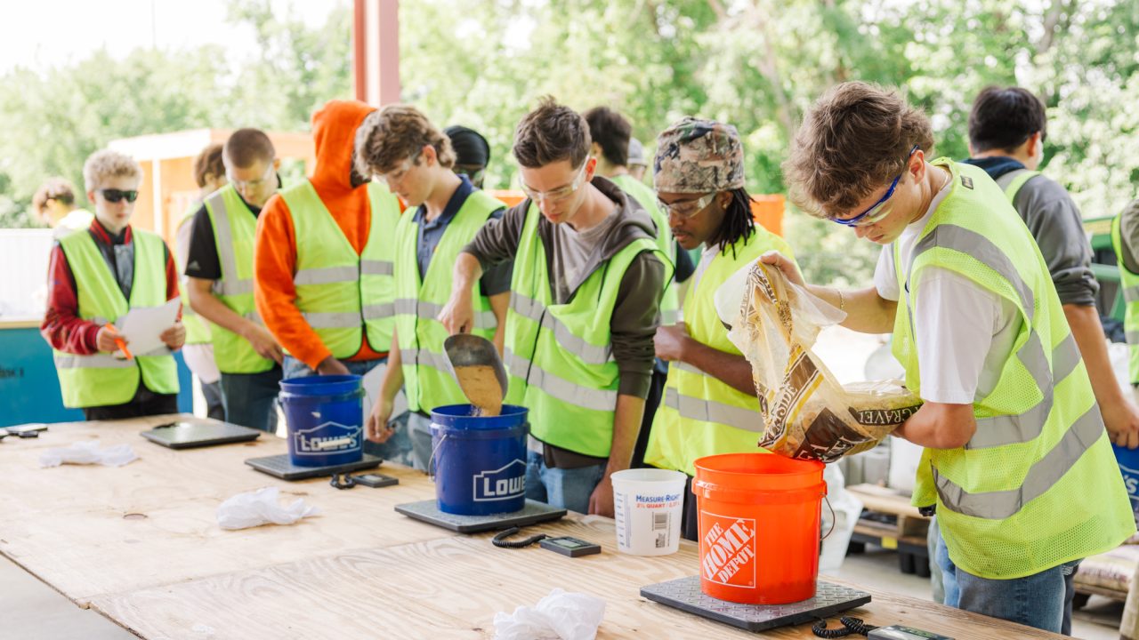 A group of people wearing yellow safety vests measure and pour materials into buckets on a wooden table during an outdoor workshop.