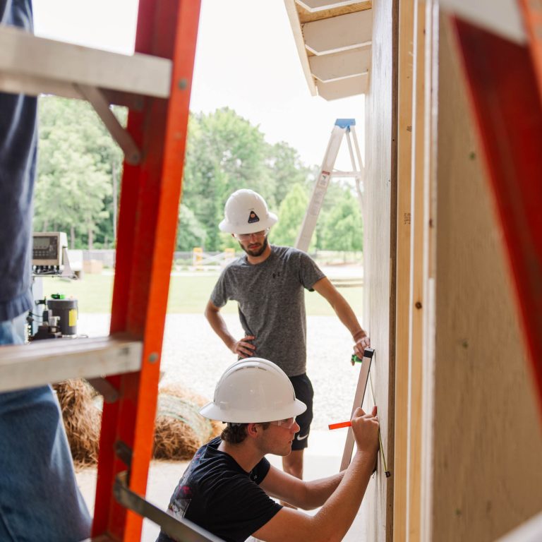 Two people wearing white hard hats work on the exterior of a building with ladders visible in the foreground and background.