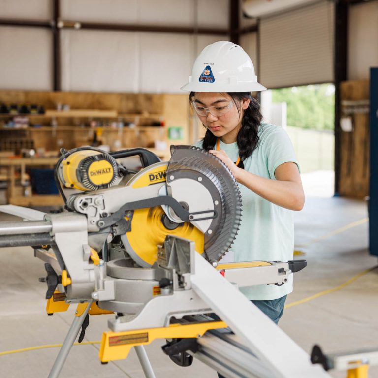 A person wearing safety goggles and a hard hat operates a DeWalt miter saw in a workshop. Shelves with tools and materials are visible in the background.