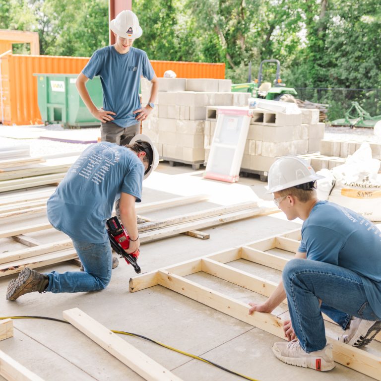 Three people in hard hats and blue shirts assemble wooden frames at a construction site; one uses a nail gun while another holds a board in place.