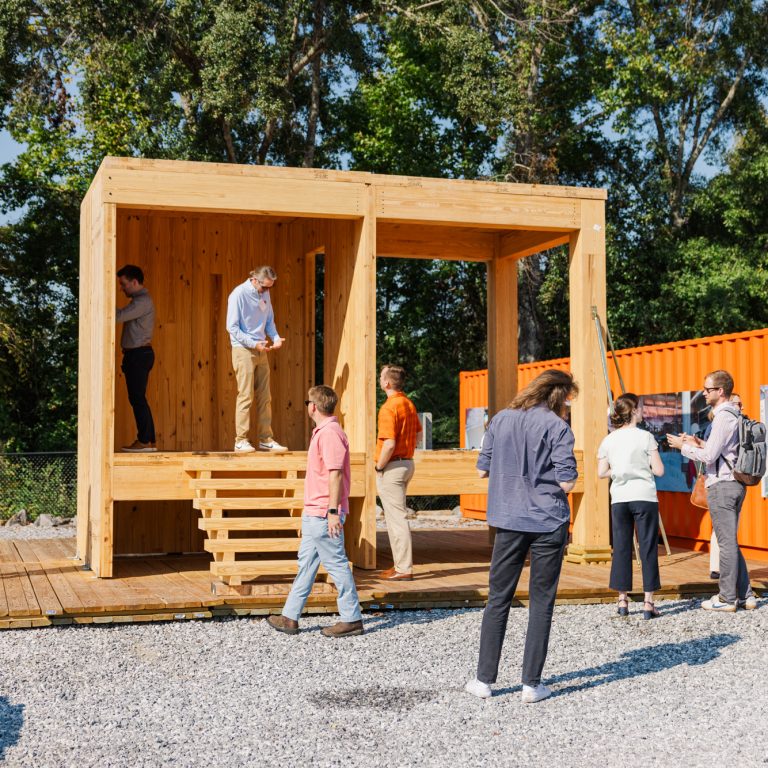 A group of people gathers around and on a small modern wooden structure outdoors, with orange shipping containers and trees in the background.