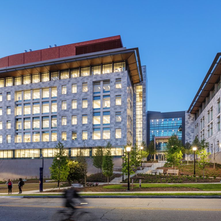 Modern university buildings with large windows at dusk, a few people walking, and a cyclist passing by on the street in the foreground.