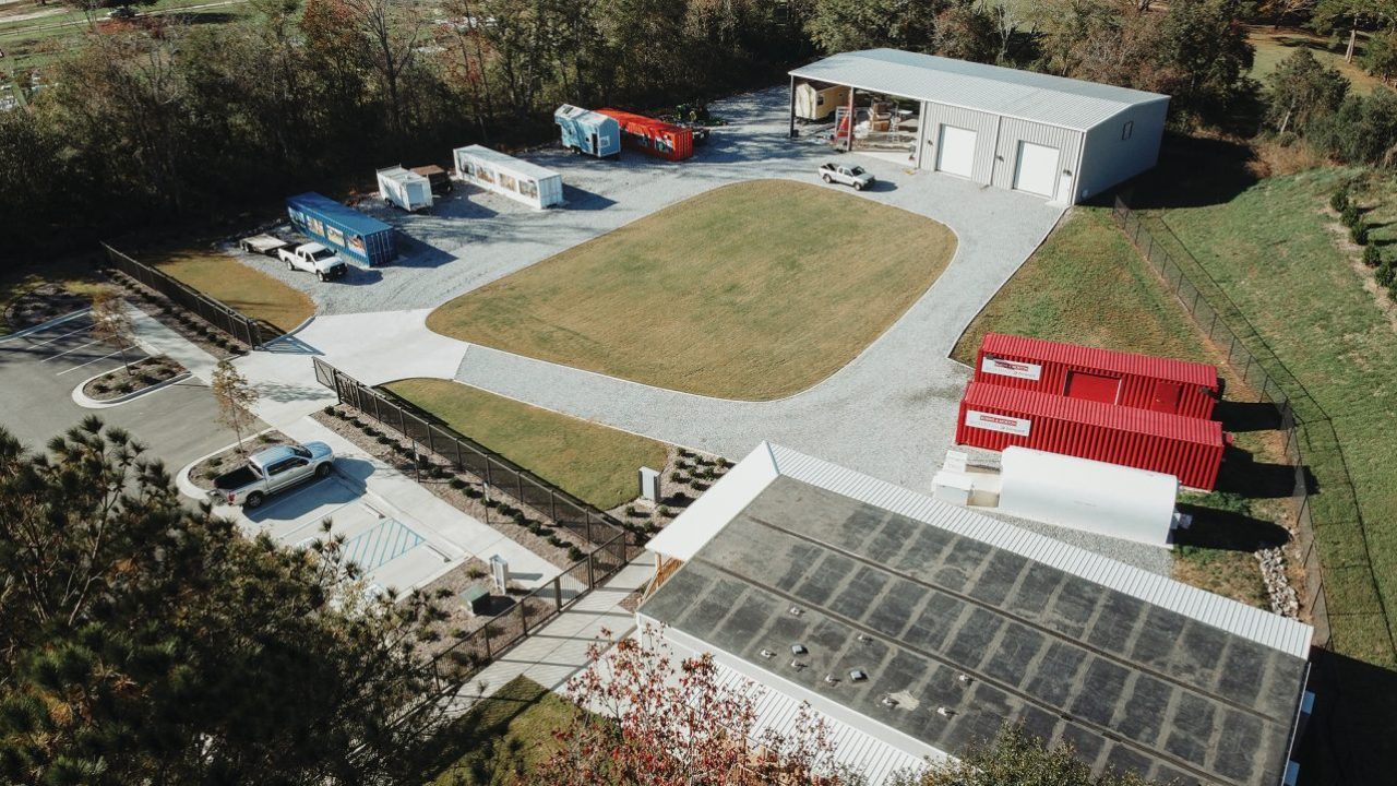 Aerial view of an industrial lot with several shipping containers, two large buildings, vehicles, and a central grassy area surrounded by gravel paths.