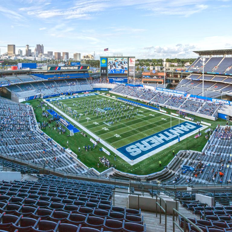 Wide view of a football stadium with a game in progress, partially filled stands, and city skyline visible in the background.