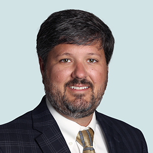 A man with dark hair and a beard, wearing a plaid suit jacket, white shirt, and striped tie, poses in front of a light blue background.