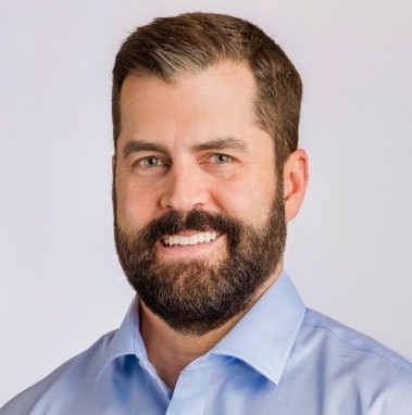A man with short brown hair and a beard, wearing a light blue collared shirt, is smiling against a plain light background.