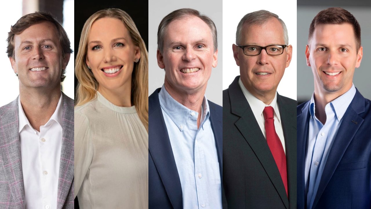 Five professionally dressed adults, three men and two women, are standing side by side and smiling in front of a plain background.