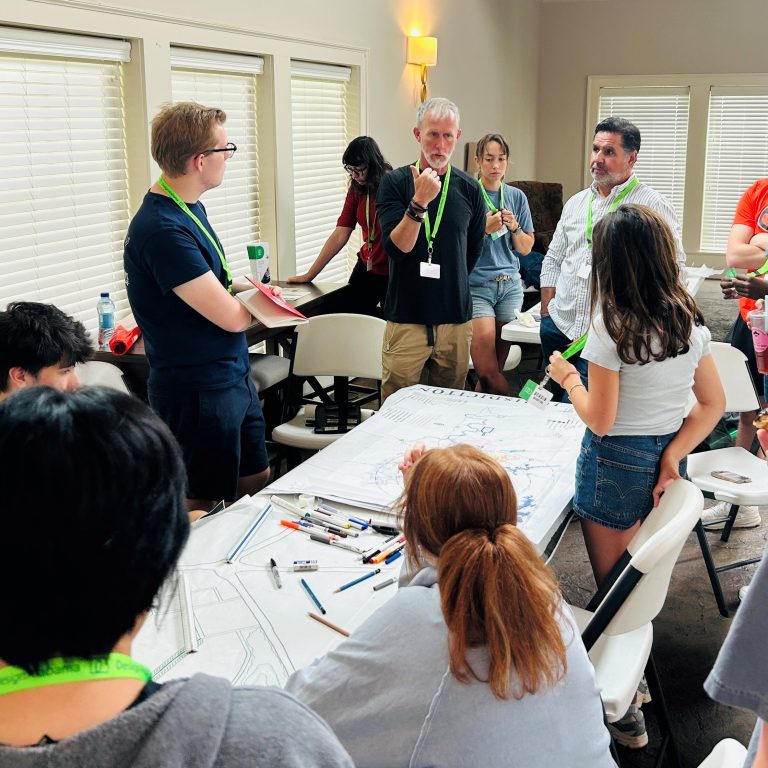 A group of people gathers around a table covered with papers and markers, actively discussing and collaborating in a well-lit room.