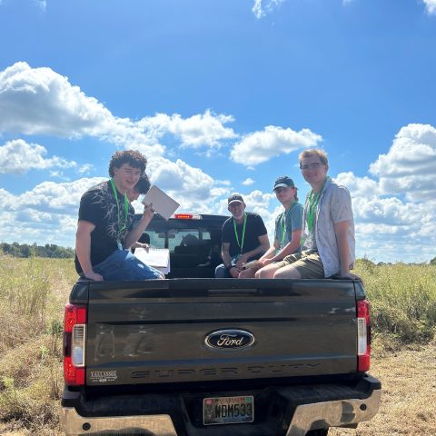 Four people with green lanyards sit on the tailgate of a Ford Super Duty truck in a grassy field under a blue sky with clouds.