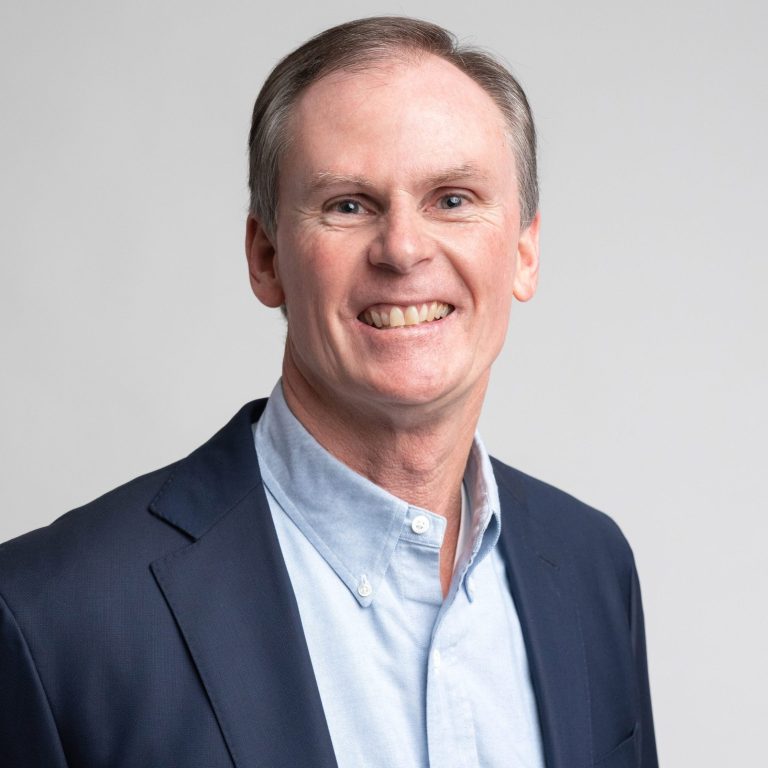 A middle-aged man with short brown hair, wearing a light blue shirt and a navy blazer, smiling at the camera against a plain light background.