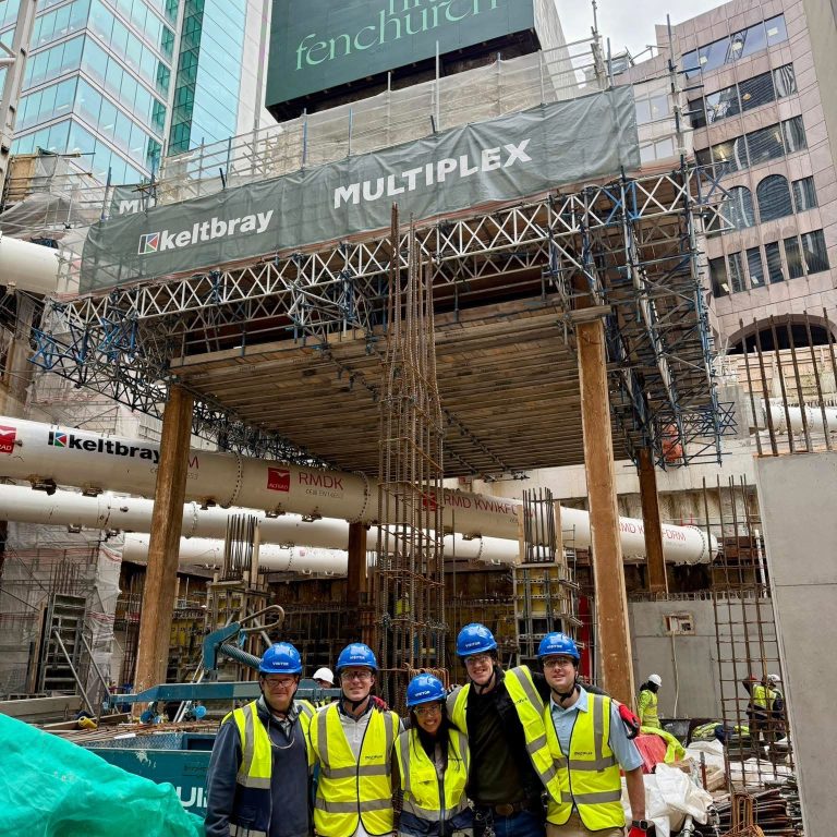 Five construction workers in safety gear stand together in front of a building site scaffolded with signs reading fifty fenchurch, keltbray, and MULTIPLEX in an urban area.